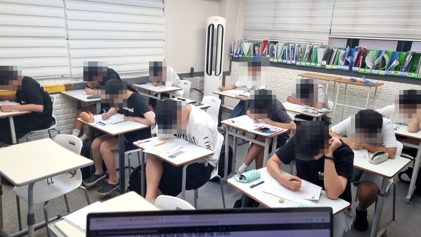 넓은 강의실에서 여러 학생이 동시에 시험지를 풀고 있는 장면
